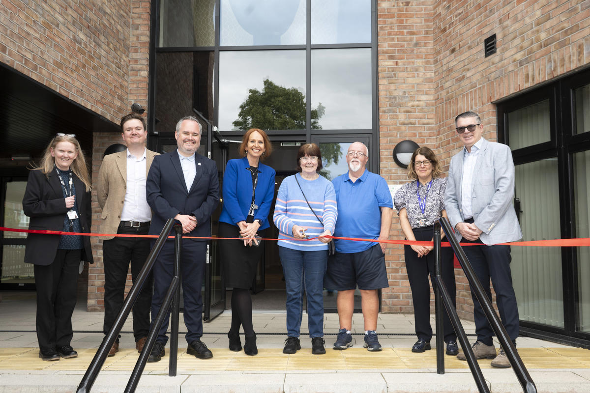 Collection of people in front of building with a red ribbon about to be cut