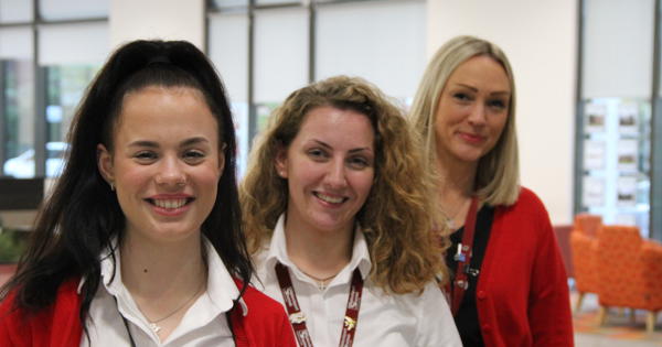 3 women lined up in white shirts and red jackets in a reception of an building
