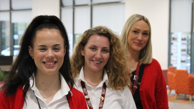 3 women lined up in white shirts and red jackets in a reception of an building